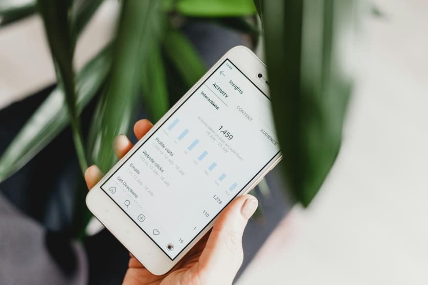 Hand holding a white smartphone displaying a business insights page with engagement metrics and blue bar charts, photographed against green plant leaves