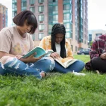Three Black college students sitting on the grass outdoors and studying from books.