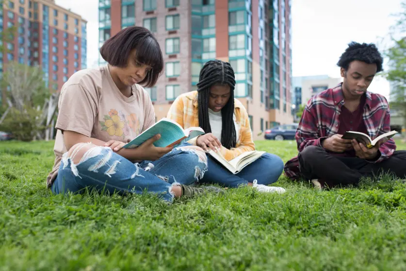 Three Black college students sitting on the grass outdoors and studying from books.